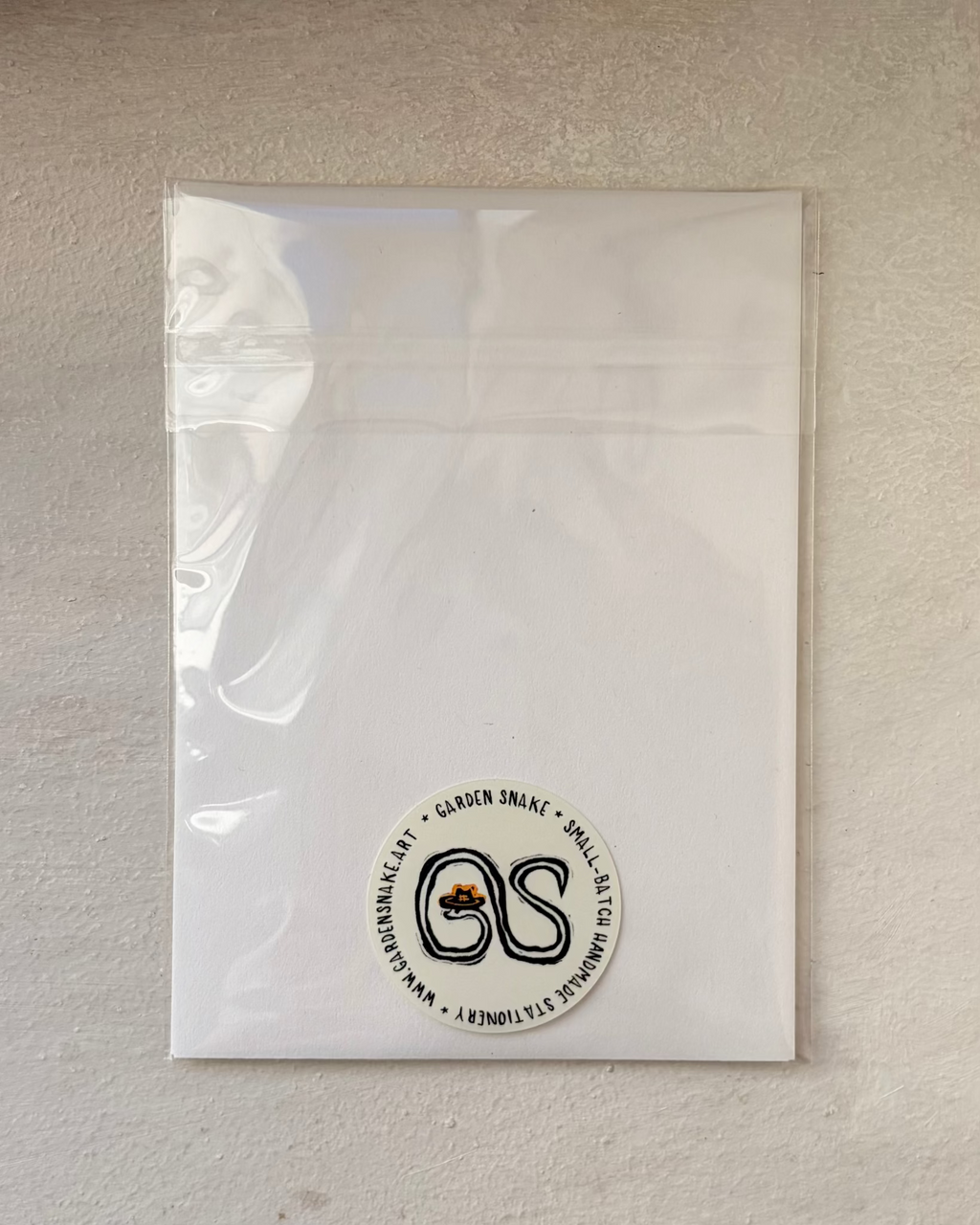 A clear plastic resealable pouch containing a white insert or backing card, photographed against a textured off-white wall. A circular black-and-white logo sticker is visible near the bottom center of the pouch.