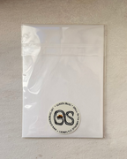 A clear plastic resealable pouch containing a white insert or backing card, photographed against a textured off-white wall. A circular black-and-white logo sticker is visible near the bottom center of the pouch.