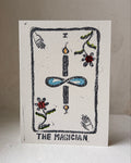 A square white art print card titled 'THE MAGICIAN' featuring a hand-drawn tarot card design with a black outlined border, a central blue infinity symbol, and small colorful floral and hand illustrations, photographed standing upright against a neutral beige background.