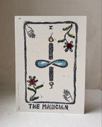 A square white art print card titled 'THE MAGICIAN' featuring a hand-drawn tarot card design with a black outlined border, a central blue infinity symbol, and small colorful floral and hand illustrations, photographed standing upright against a neutral beige background.