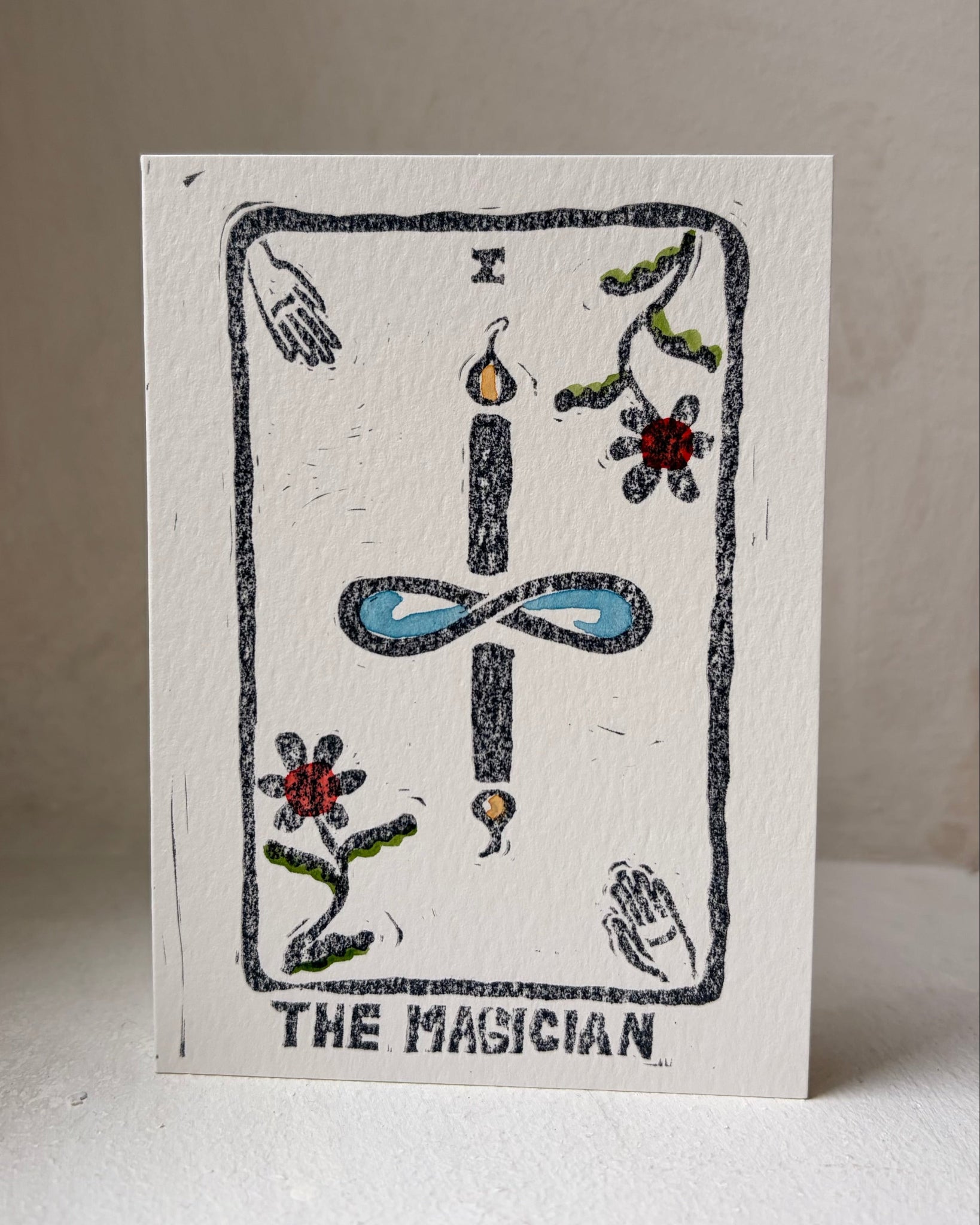 A square white art print card titled 'THE MAGICIAN' featuring a hand-drawn tarot card design with a black outlined border, a central blue infinity symbol, and small colorful floral and hand illustrations, photographed standing upright against a neutral beige background.