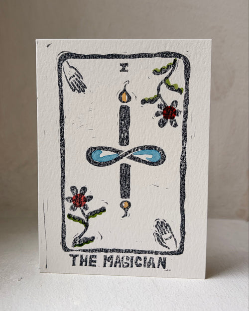 A square white art print card titled 'THE MAGICIAN' featuring a hand-drawn tarot card design with a black outlined border, a central blue infinity symbol, and small colorful floral and hand illustrations, photographed standing upright against a neutral beige background.