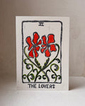 A square white card featuring a tarot-style illustration labeled 'THE LOVERS,' with two red flowers, green vines, and black border details, photographed standing against a plain light background.