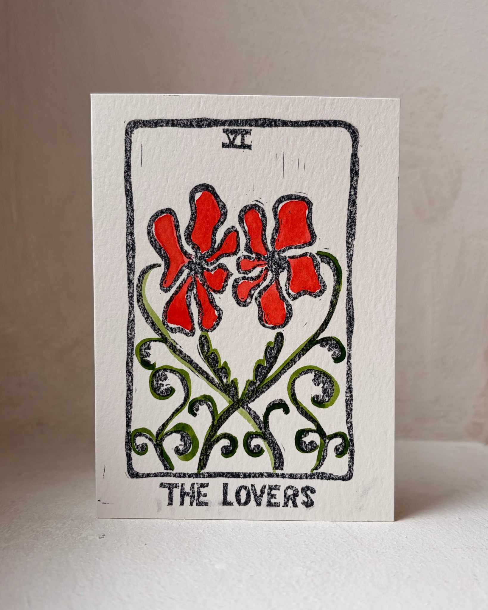 A square white card featuring a tarot-style illustration labeled 'THE LOVERS,' with two red flowers, green vines, and black border details, photographed standing against a plain light background.