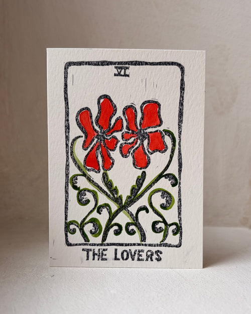 A square white card featuring a tarot-style illustration labeled 'THE LOVERS,' with two red flowers, green vines, and black border details, photographed standing against a plain light background.