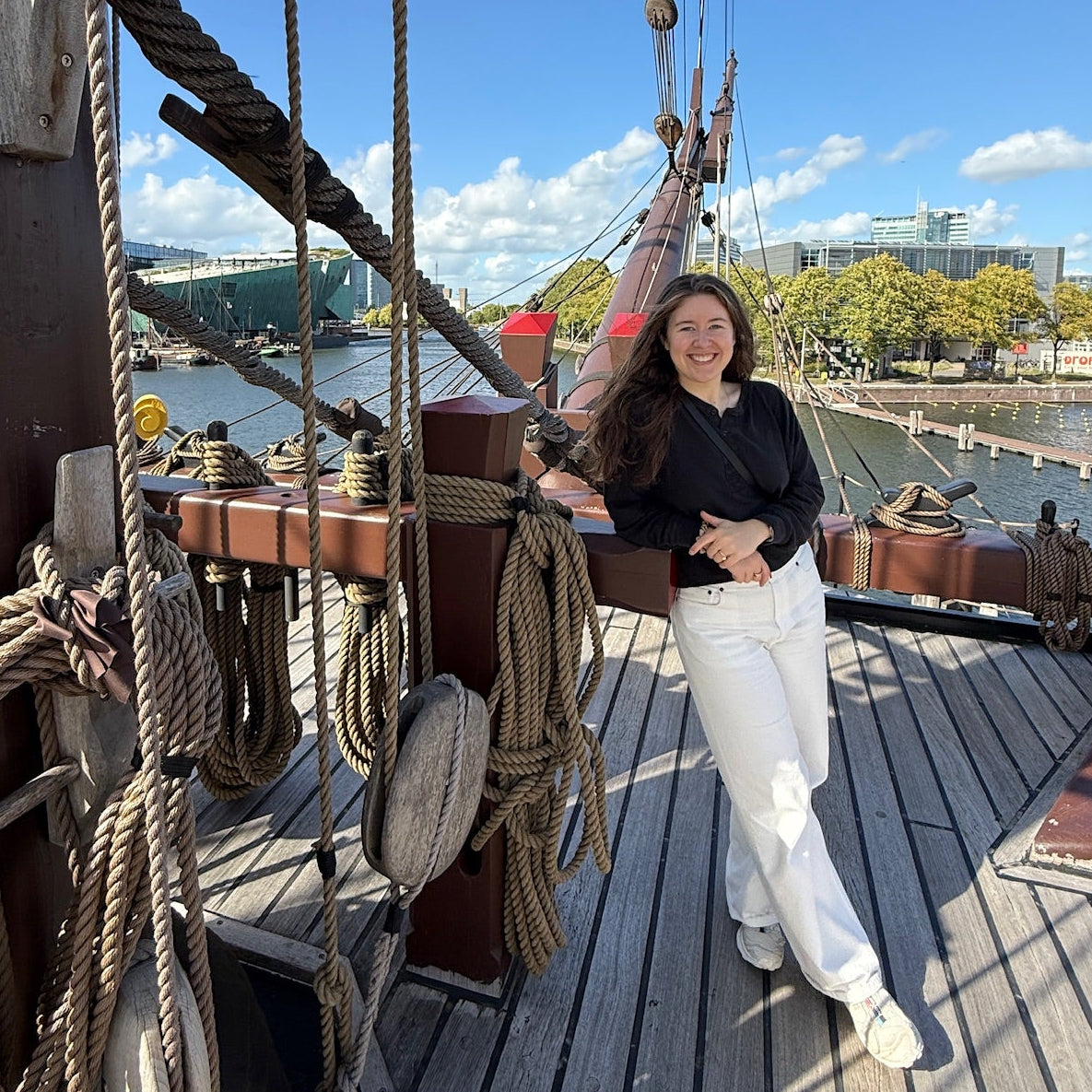 A woman posing on the wooden deck of a sailing ship replica, wearing a black long-sleeve top and white pants, with ropes, rigging, and a blue sky background.