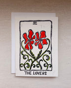 A square art print card featuring a tarot-style illustration labeled 'THE LOVERS' with red flowers, green vines, and black border details, beneath, an envelope.