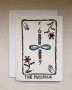 A tarot card art print card titled 'THE MAGICIAN' displayed on a textured beige wall, featuring a black ink linocut style illustration with blue, red, and green accents on an off-white cardstock. An envelope beneath.