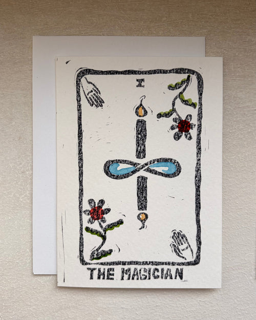 A tarot card art print card titled 'THE MAGICIAN' displayed on a textured beige wall, featuring a black ink linocut style illustration with blue, red, and green accents on an off-white cardstock. An envelope beneath.
