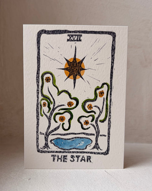 A square art print card titled 'THE STAR' featuring a tarot-style illustration with a black star, green vines, orange flowers, and a blue pool on an off-white background, photographed standing upright against a neutral beige backdrop.