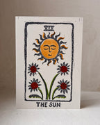 A square art print card featuring a tarot-style illustration labeled 'THE SUN,' with a yellow sun face, black rays, red flowers, and green stems on an off-white background, photographed against a neutral light beige wall and surface.